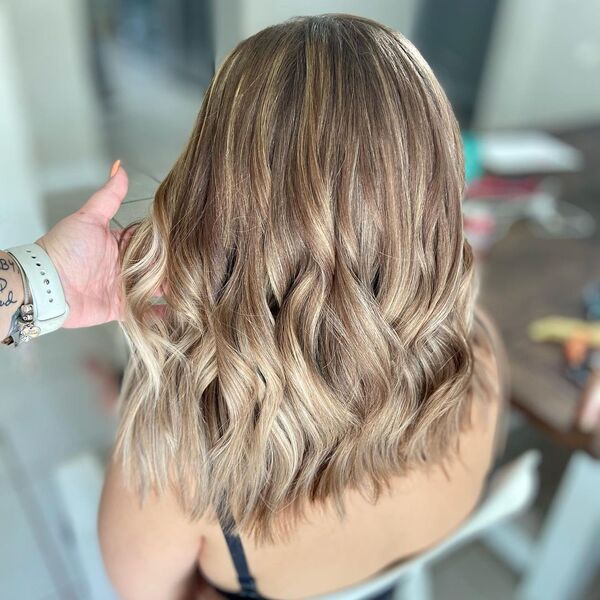 Dimensional Blonde Balayage - a woman sitting and another woman with orange nails is holding her hair